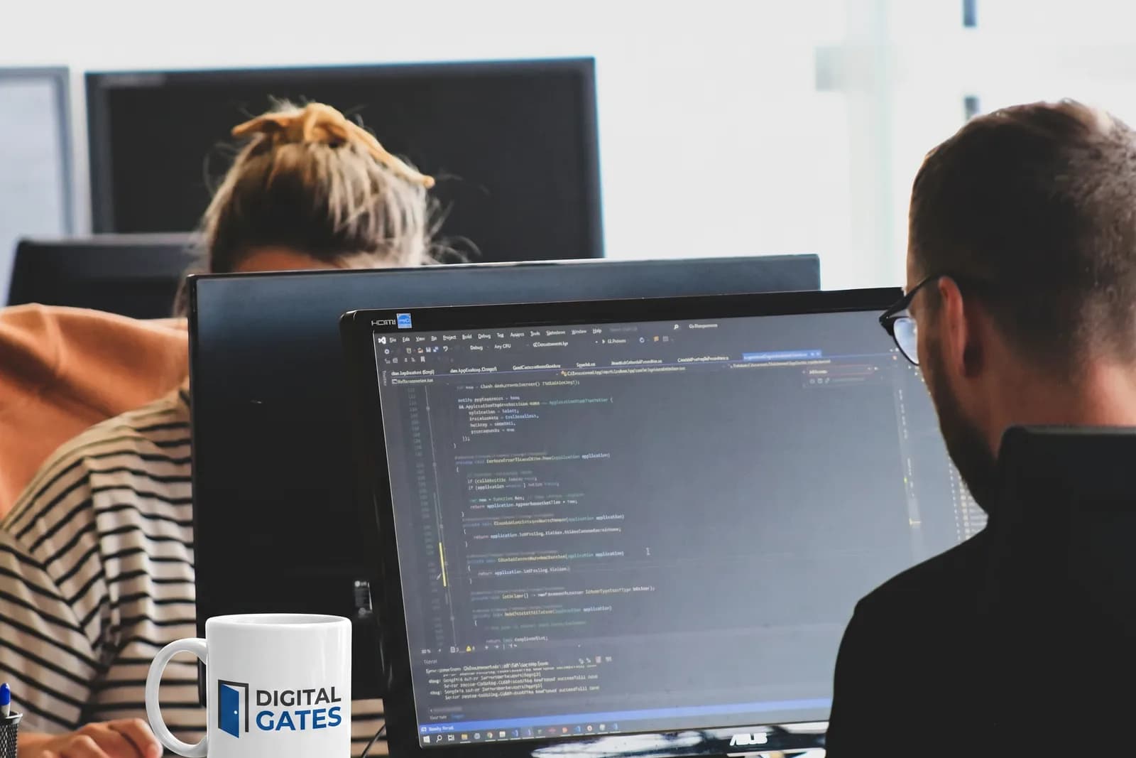Two Digital Gates engineers reviewing .NET code on a desktop monitor, a branded mug on the desk beside them